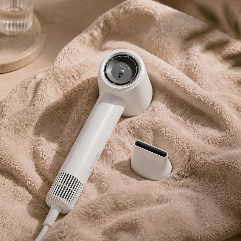 A white cordless hair dryer with an attached nozzle lies on a beige towel, with a matching white charging or accessory piece beside it. The scene is styled with warm neutral tones and soft shadows.