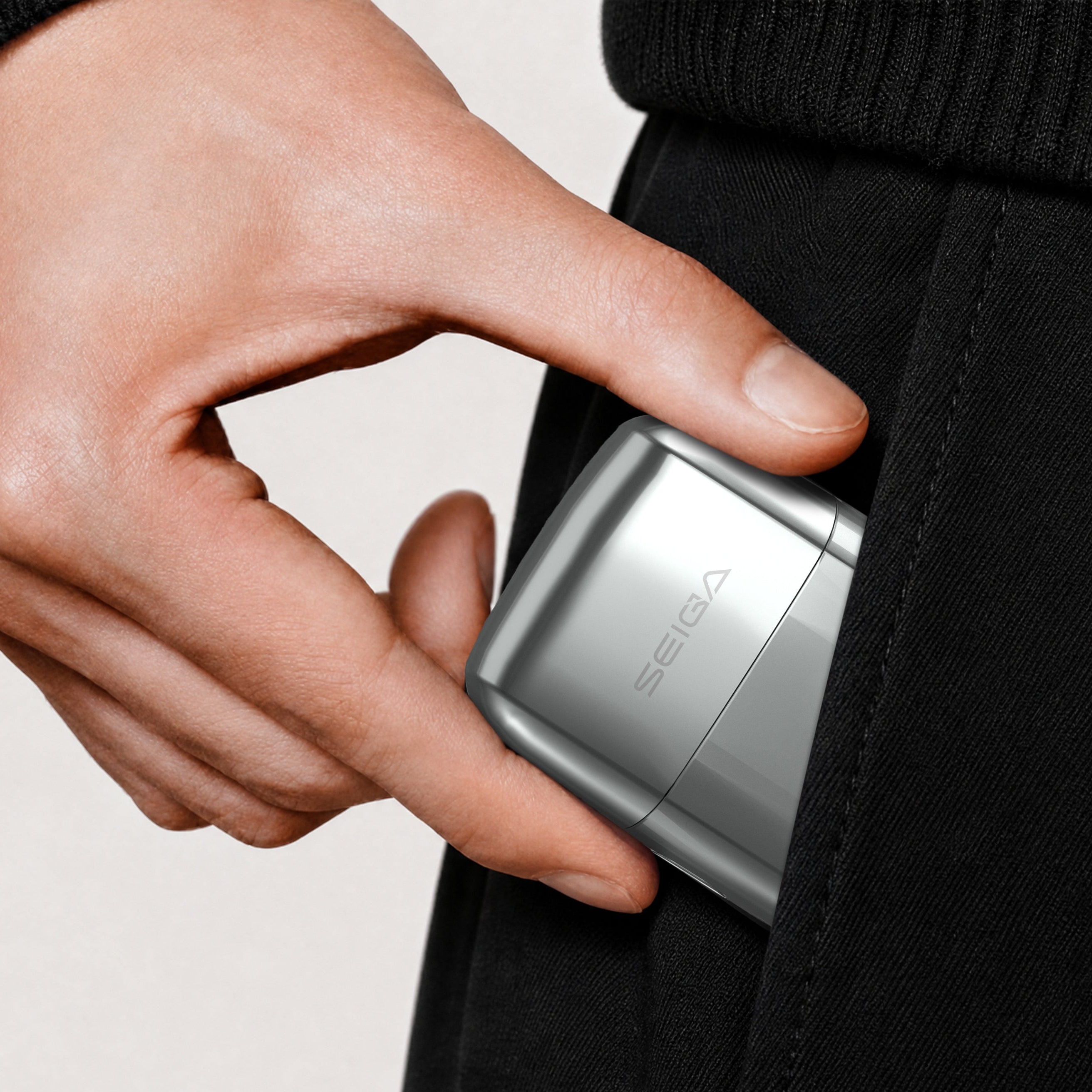 A close-up of a hand sliding a slim silver Seica digital camera into a black pocket, with a light gray background.