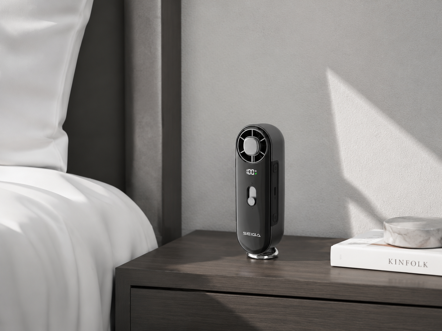 A sleek black portable fan with a silver circular grille and a small green LED indicator, standing on a metallic base on a dark wood bedside table next to a bed.