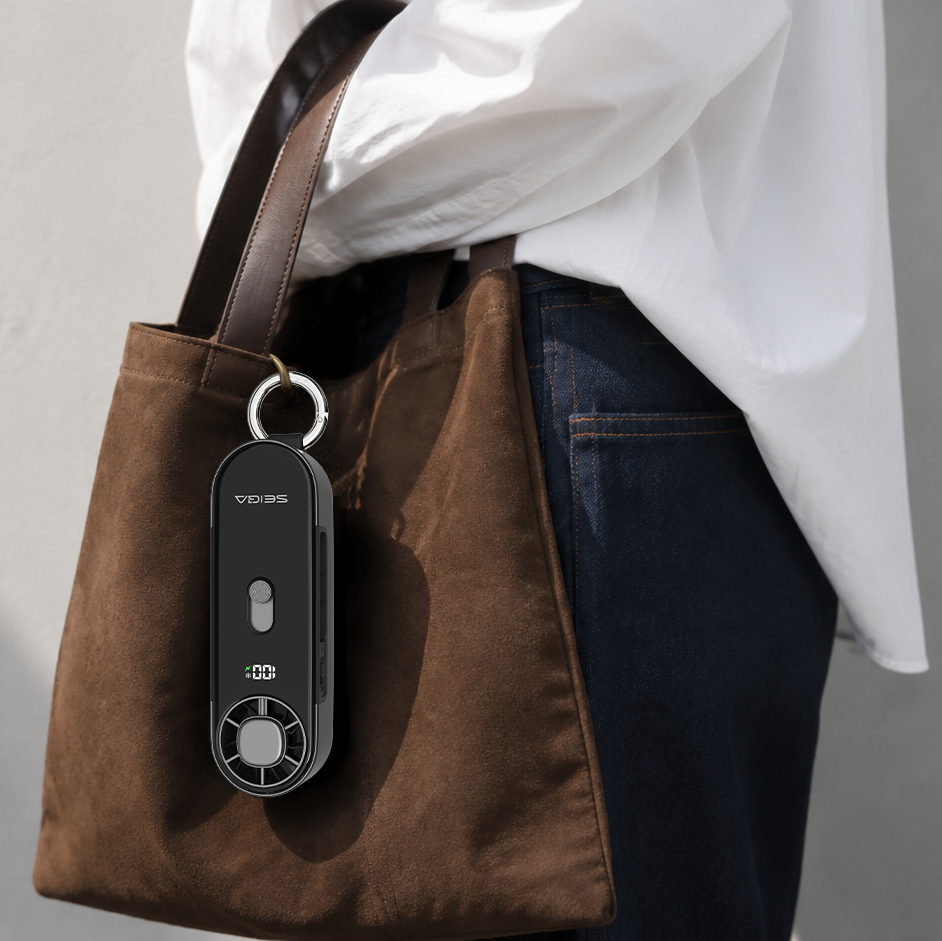 A person wearing a white shirt and dark jeans carries a brown suede tote bag against a light gray wall. A black rectangular keychain or remote-style accessory with a silver ring is clipped to the bag.