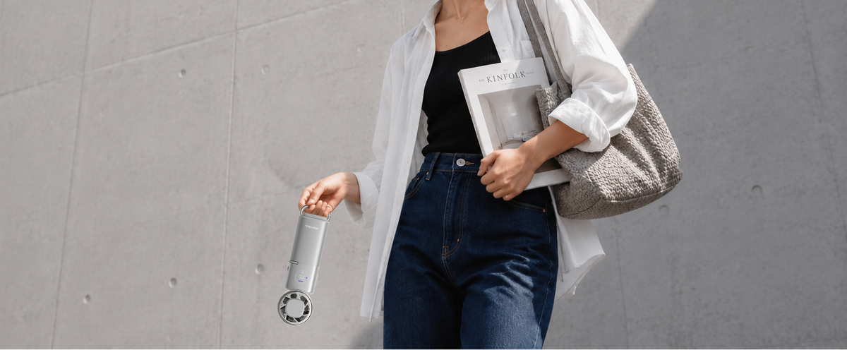 A woman walking outdoors against a concrete wall, holding a white handheld fan and carrying a white shirt, black top, blue jeans, and a gray woven tote bag.