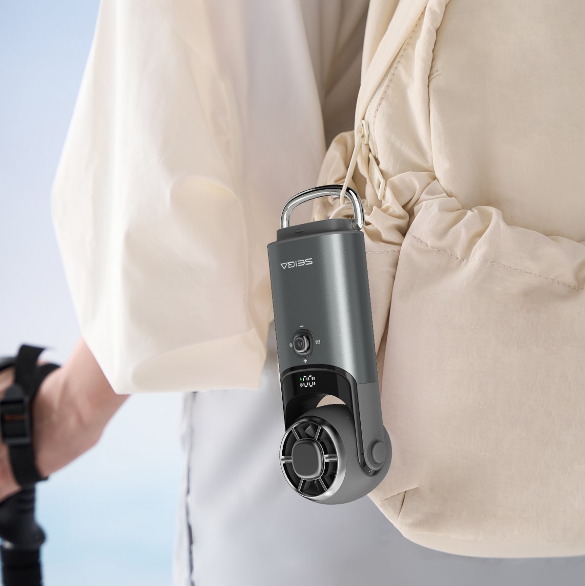 A close-up lifestyle shot of a person wearing a cream-colored hooded jacket, with a small gray VEVIMS handheld fan clipped to the side of the jacket.