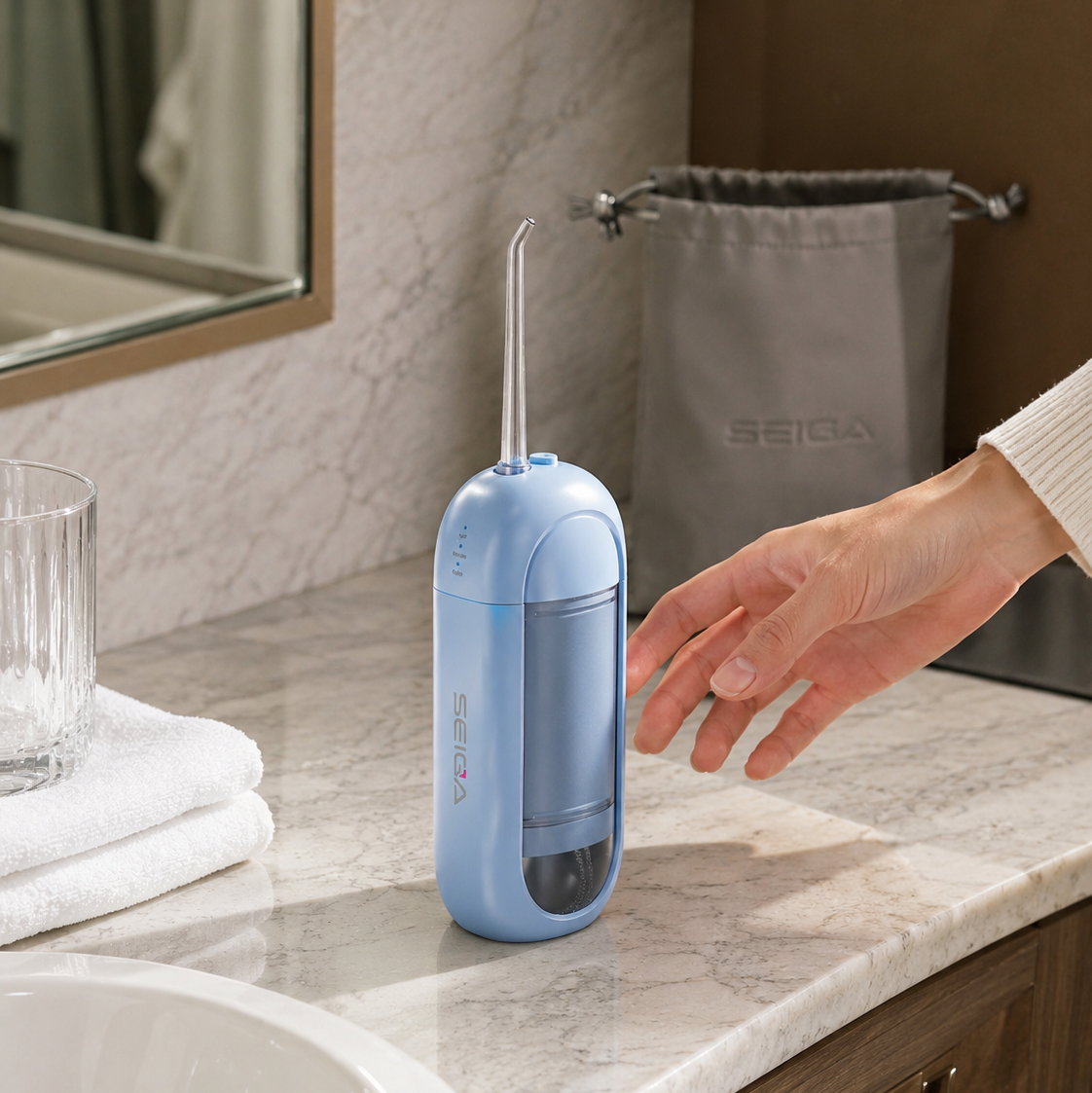A light blue oral irrigator placed on a marble bathroom counter next to a sink, with a person's hand reaching toward it. A gray drawstring bag with the brand name SEIDAI hangs in the background.