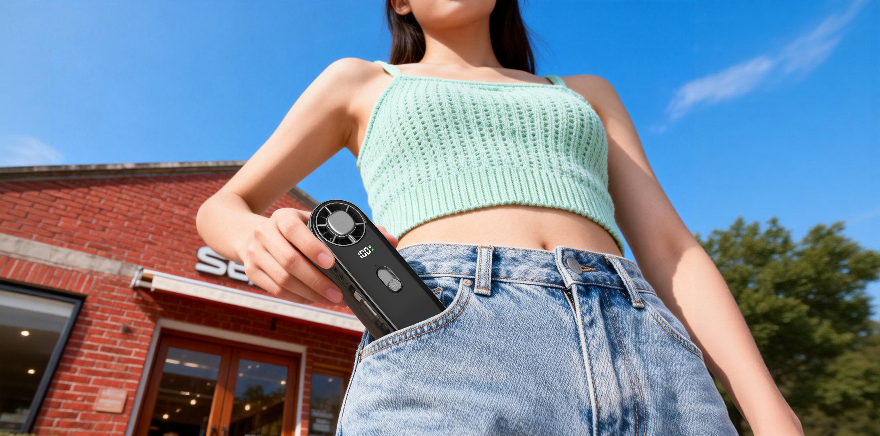 A woman outdoors in a mint green knit crop top and blue jeans pockets a black handheld circular device with a visible LED light and small printed logo. A brick storefront and blue sky are in the background.