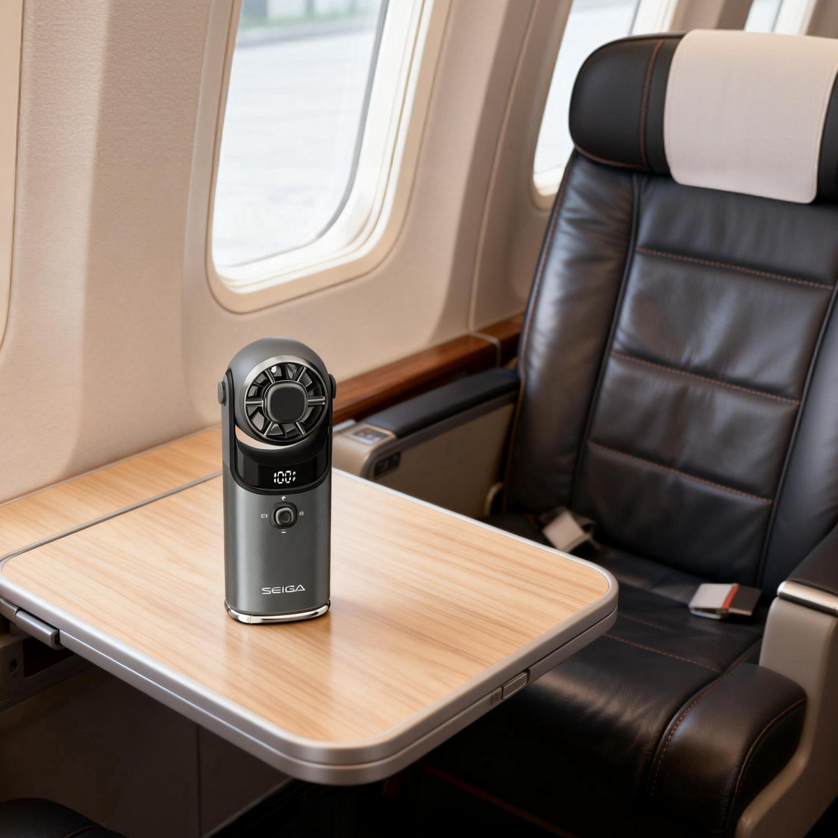 A compact black handheld fan with a circular mesh grille sits on a light wood-trimmed fold-out table inside an airplane cabin next to a black leather passenger seat.