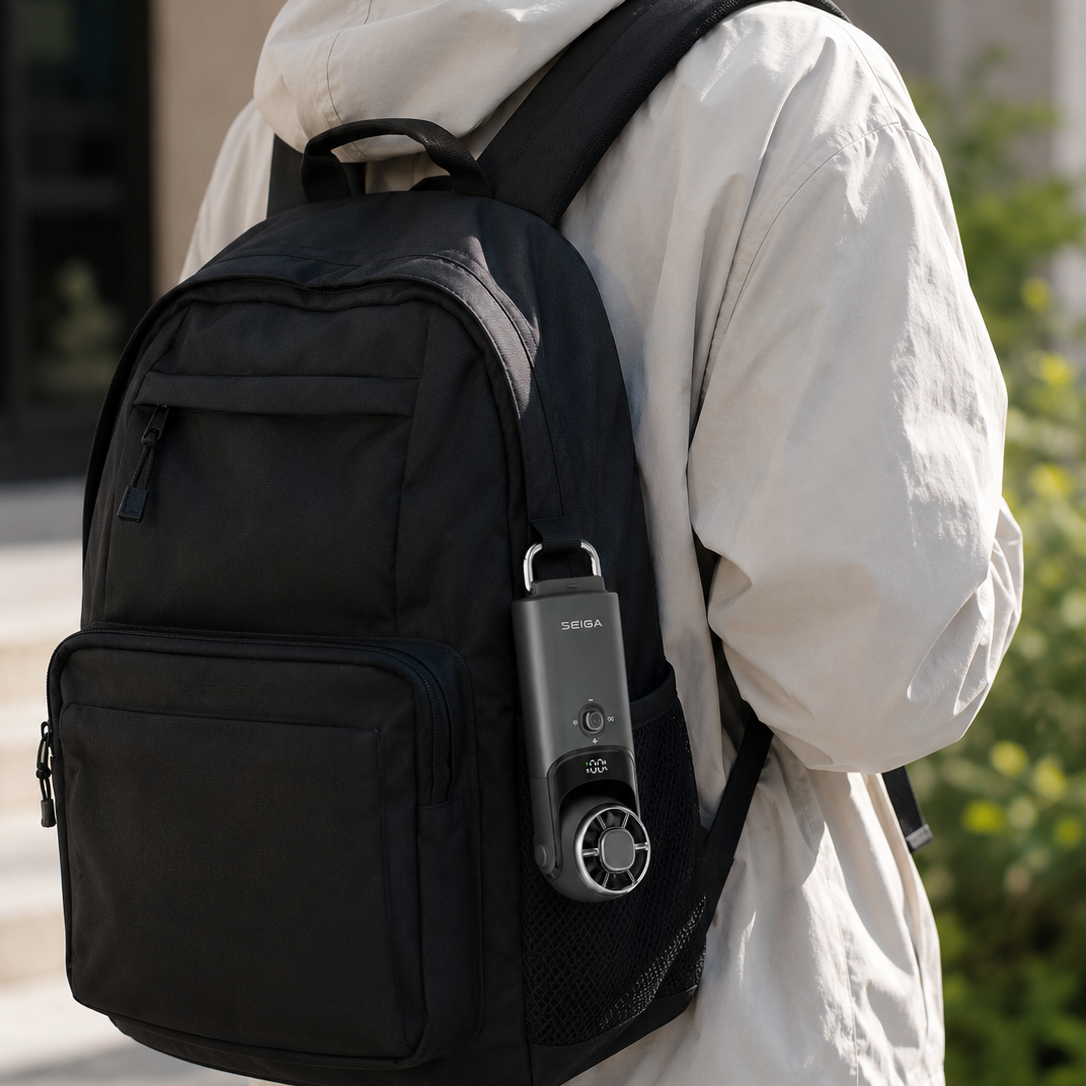 A person wearing a light beige hooded jacket is shown from the back, carrying a black backpack outdoors. The backpack has a front zip pocket, a side mesh pocket holding a silver electronic device, and padded shoulder straps.