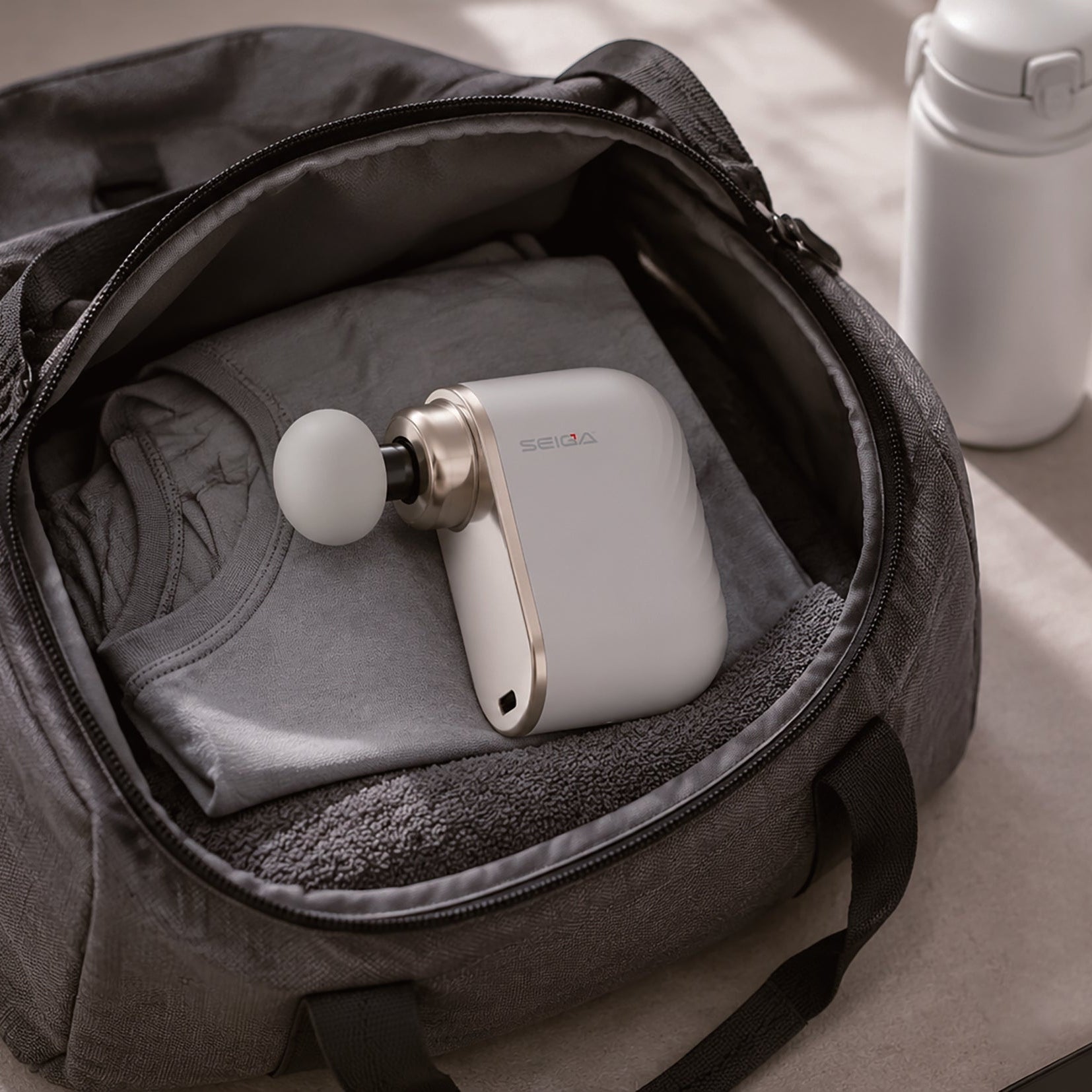 A white and gold handheld massage gun with a white charging case sits inside an open gray duffel bag, surrounded by fitness items including a white bottle, a rolled gray mat, a white sneaker, and a smartphone on a beige surface.