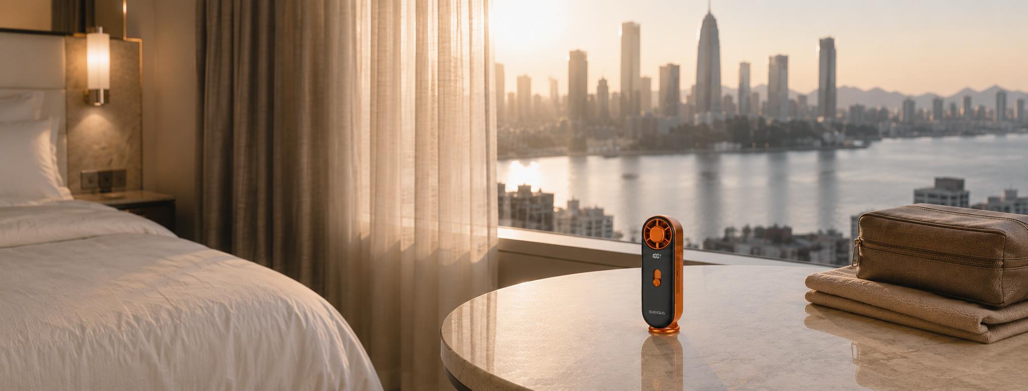 A small orange and black handheld device stands on a round marble bedside table in a modern hotel room. A bed with white linens is visible on the left, and a large window overlooks a waterfront city skyline at sunset.