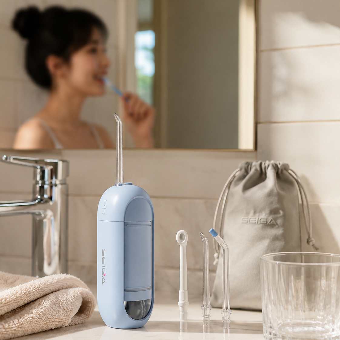 A light blue oral irrigator with three interchangeable nozzles, a beige drawstring pouch, and a clear glass cup on a bathroom counter, with a woman brushing her teeth visible in the mirror reflection.