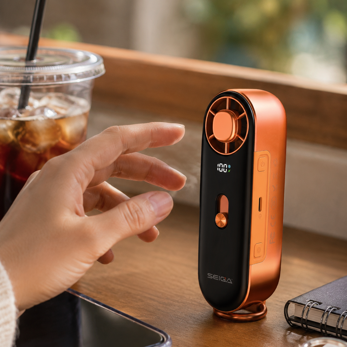 A handheld portable fan in a matte black and metallic copper-orange finish, standing on a wooden table next to a person's hand, a drink, a smartphone, and keys.