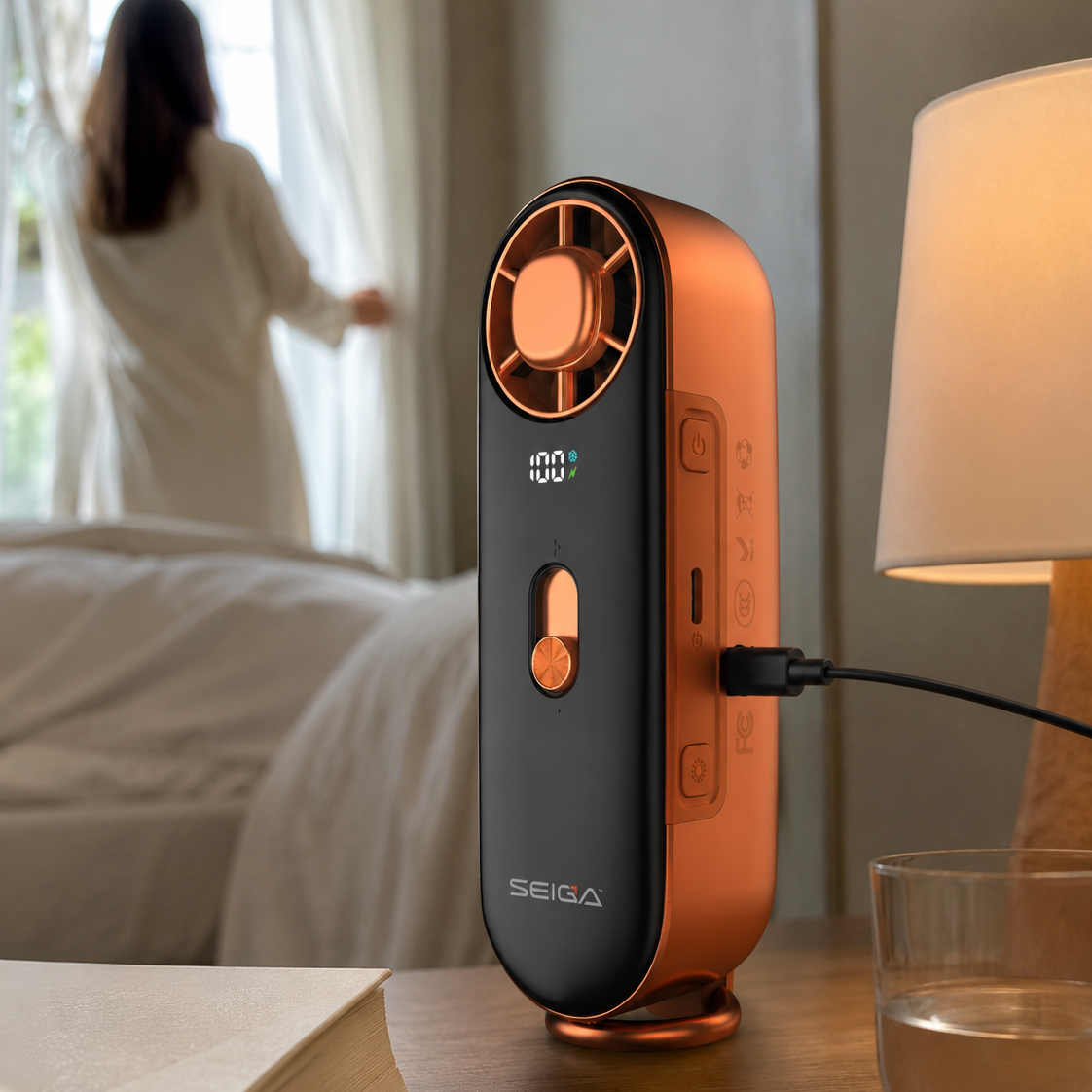 A SEIGAA portable fan and humidifier device in a matte black finish with metallic copper accents, standing on a wooden bedside table next to a lit lamp and a glass of water, with a blurred bedroom scene in the background.