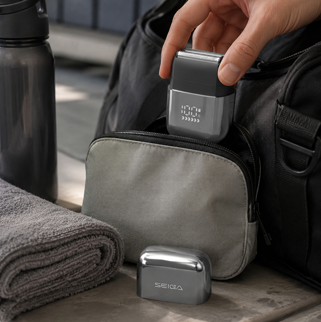 A hand inserts a gray rectangular electronic device into a zippered gray pouch beside a black duffel bag, with a folded towel and a dark bottle nearby. A smaller metallic SEIDA device sits in front on the surface.